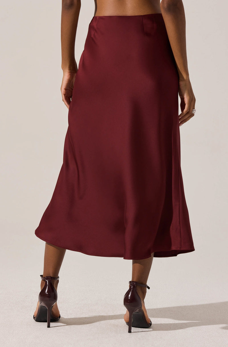 A woman is wearing a long, high-waisted, satin skirt in a deep burgundy color that reaches mid-calf length. The skirt has a smooth texture and a subtle sheen, with a clean, simple design. She is also wearing dark brown high-heeled shoes with ankle straps.