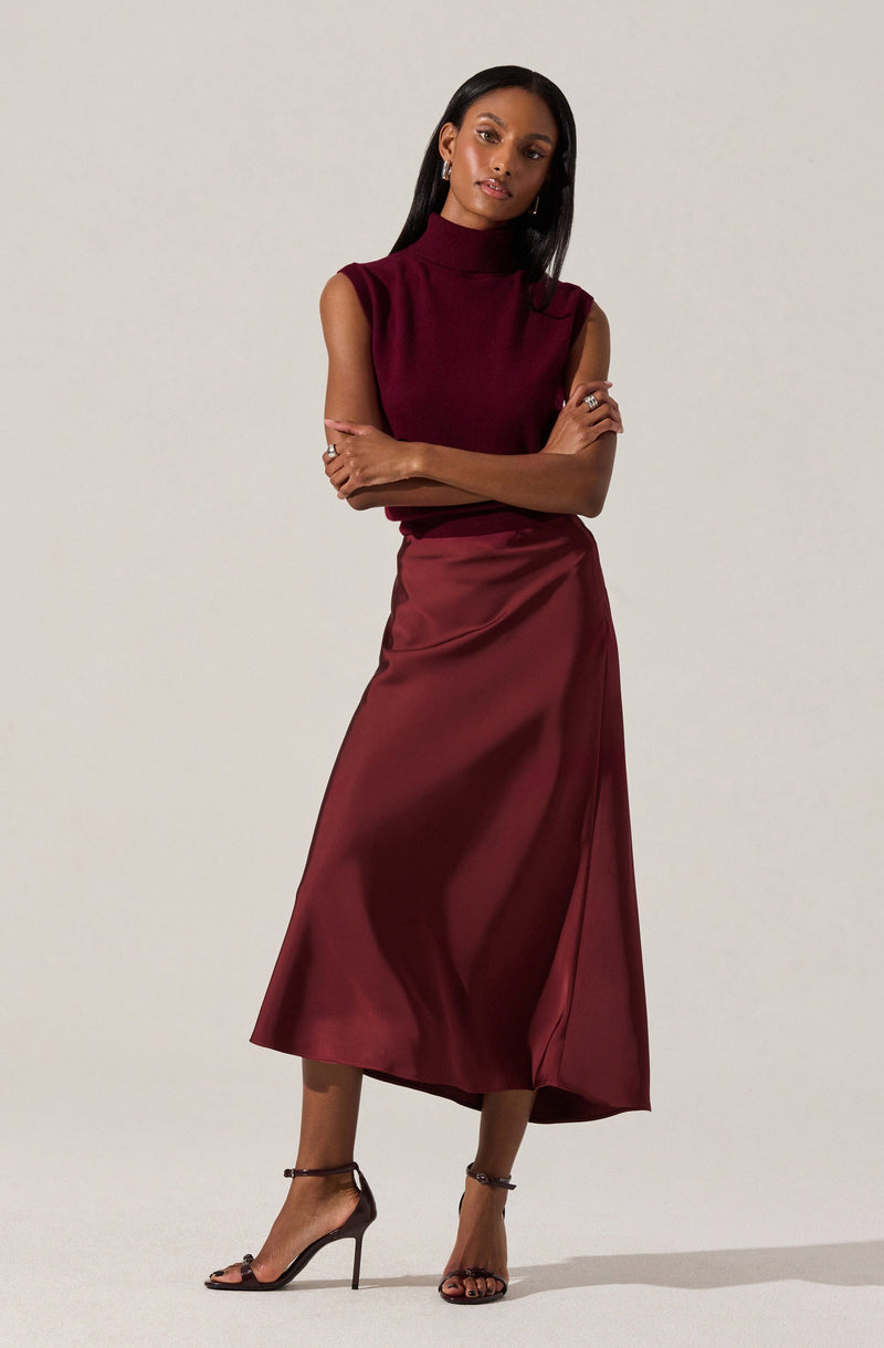 A woman is wearing a sleeveless, high-neck, knit top in a deep burgundy color. She pairs it with a matching burgundy satin midi skirt that has a slight sheen and a slit on the side. She completes her outfit with black high-heeled sandals with ankle straps.