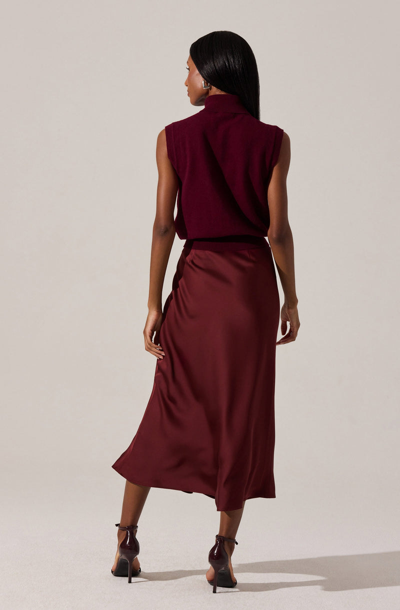 A woman is wearing a sleeveless, high-neck burgundy top with shoulder padding, paired with a matching satin midi skirt that has a slight sheen and a slit at the back. She is also wearing dark high-heeled shoes with ankle straps.