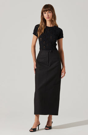 A woman is wearing a short-sleeved black lace top with intricate floral patterns, paired with a high-waisted, ankle-length black skirt that features a front button and hidden zipper. She completes her outfit with black open-toe high-heeled sandals.