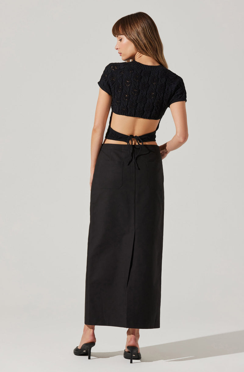 A woman is wearing a black, open-knit, short-sleeved crop top with a round neckline. The top is loosely fitted and features a textured, cable-knit pattern. She pairs it with a high-waisted, long black skirt that reaches her ankles, which has a clean, streamlined design with visible back pockets and a central seam. The skirt appears to be made of a sturdy fabric. She completes the outfit with black high-heeled mules.