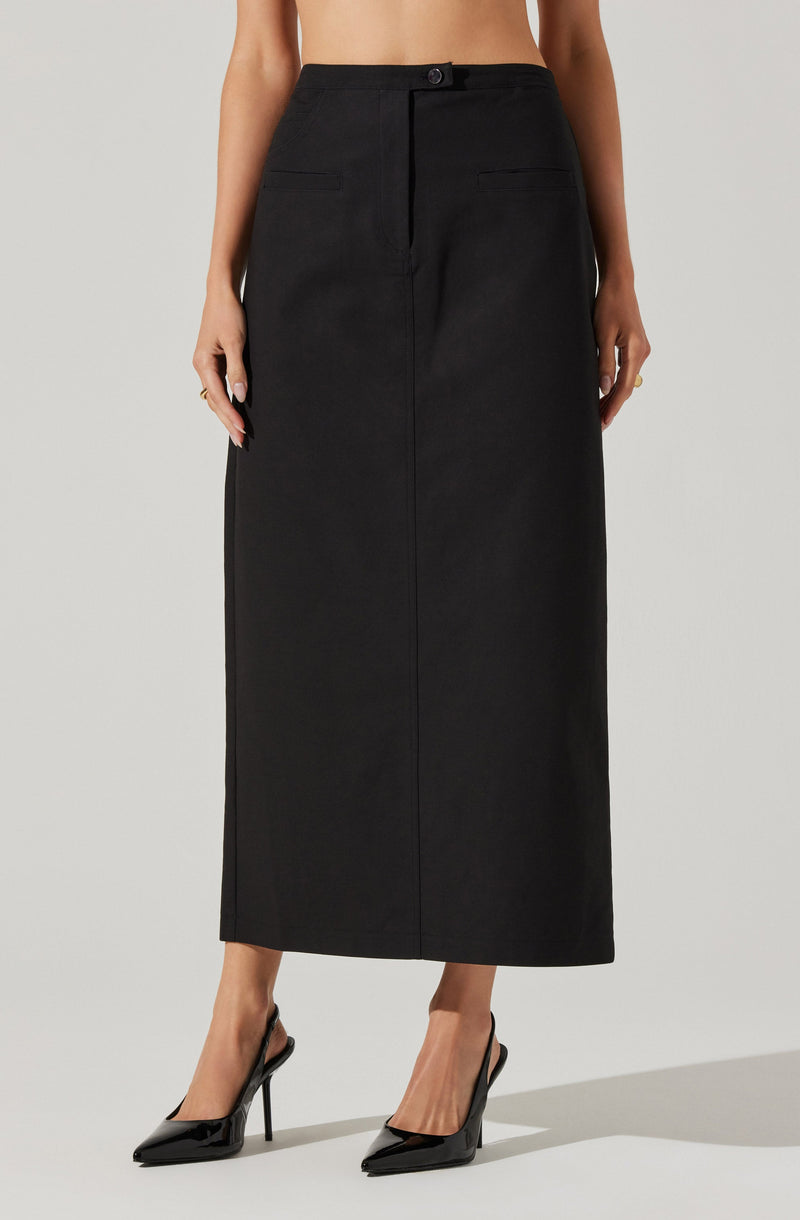 A woman is wearing a high-waisted, full-length black skirt with a straight fit and a button and zipper closure at the waistband. The skirt has two small, horizontal front pockets. She is also wearing black high-heeled pointed-toe shoes.