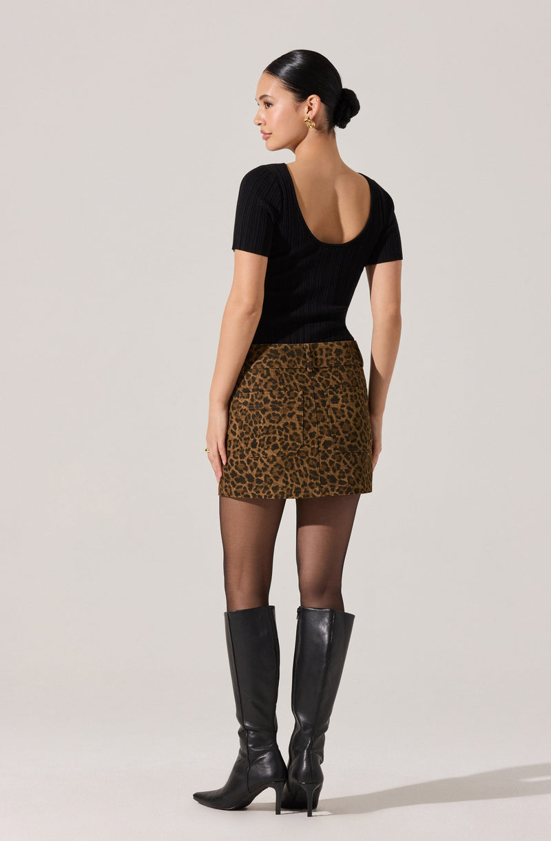 A woman is wearing a fitted black top with short sleeves and a scoop neckline, paired with a high-waisted leopard print mini skirt. She has on sheer black tights and knee-high black leather boots with pointed toes and stiletto heels.
