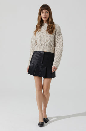 A woman is wearing a chunky, cream-colored cable knit sweater with long sleeves and a high neckline. She pairs it with a black leather mini skirt that features vertical pleats or panels. She completes the outfit with black pointed-toe shoes that have decorative embellishments on the front.