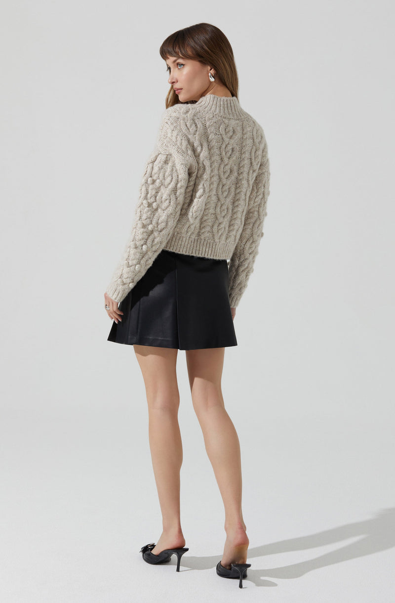 A woman is wearing a chunky, beige knit sweater with cable and bobble patterns, paired with a black leather A-line mini skirt. She is also wearing black high-heeled mules.