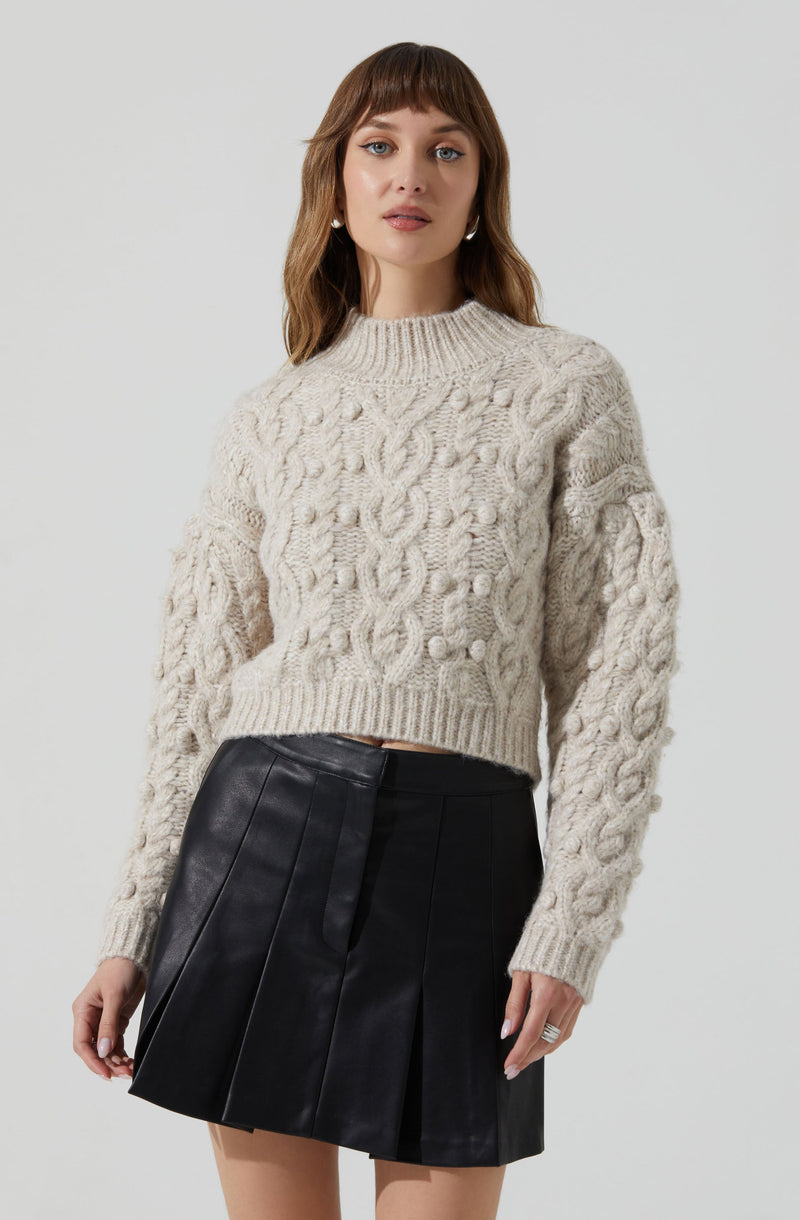 A woman is wearing an oversized, cream-colored knit sweater with a cable pattern design. She pairs it with a high-waisted black pleated leather skirt.