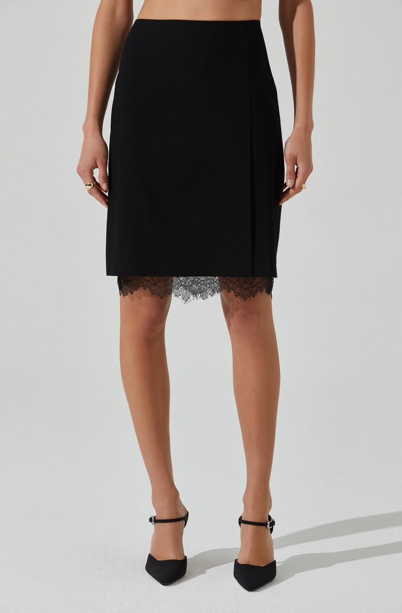 A woman is wearing a black knee-length skirt with a subtle slit on the side. The skirt features a lace trim at the hem, which is visible beneath the main fabric. She is also wearing black pointed-toe heels with ankle straps. Her hands are at her sides, and she has rings on her fingers.