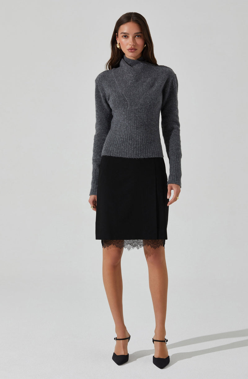 A woman is wearing a long-sleeved, high-neck, gray knit sweater with ribbed details at the cuffs and hem. She pairs it with a black knee-length skirt that features lace trim at the hem. She completes her outfit with black high-heeled shoes with ankle straps and gold earrings.