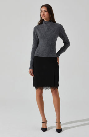 A woman is wearing a long-sleeved, high-neck, gray sweater made of a warm, textured knit fabric. She pairs it with a black skirt that falls just above the knees, featuring a lace trim at the hem. She completes her outfit with black ankle-strap heels.