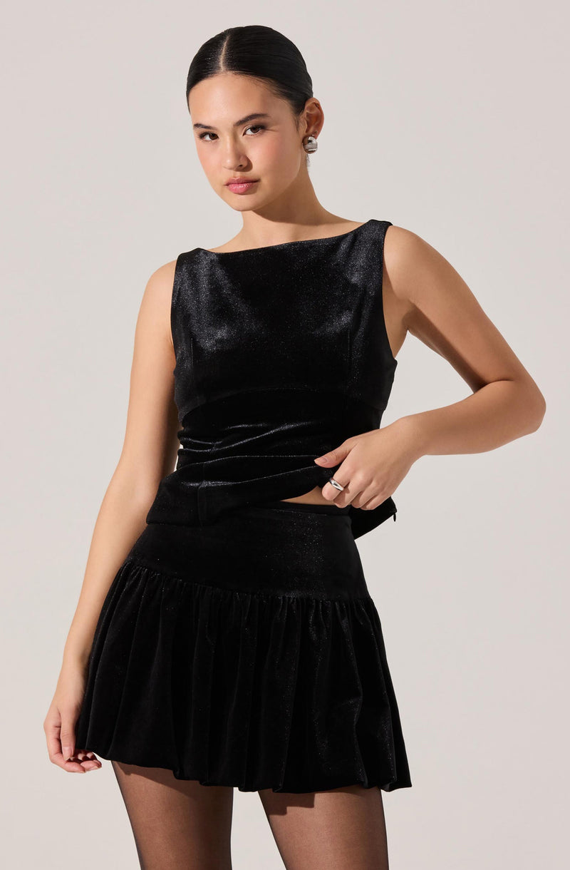 A woman is wearing a sleeveless, black, shimmery dress with a fitted bodice and a round neckline. The dress has a slightly flared, ruffled skirt made of a velvet-like material. She is also wearing sheer black tights.