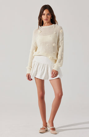 A woman is wearing a cream-colored, chunky knit sweater with a loose fit, featuring textured pom-pom details throughout. She pairs it with a white, high-waisted, ruffled mini skirt. Her accessories include gold hoop earrings, and she is wearing cream-colored, woven, low-heeled shoes with ankle straps.
