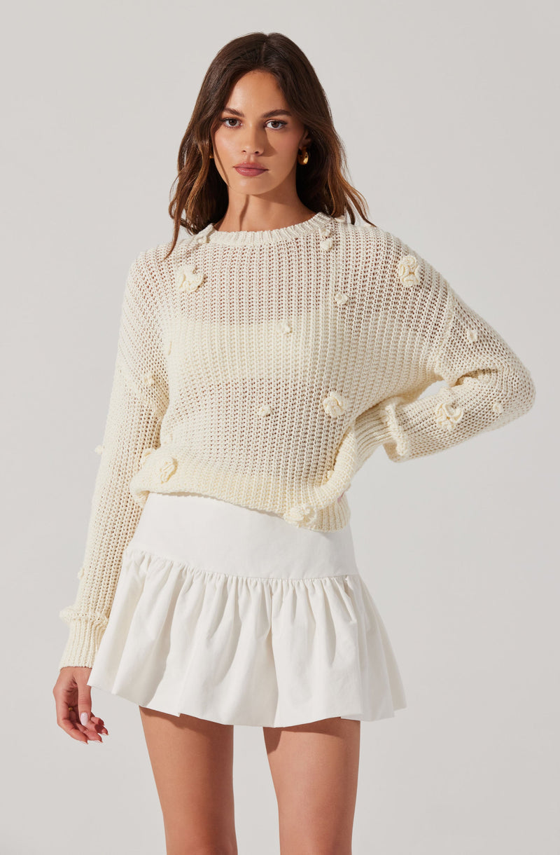 A woman is wearing a cream-colored, loose-knit sweater with small 3D floral details scattered across it. She pairs it with a white, high-waisted, ruffled mini skirt.