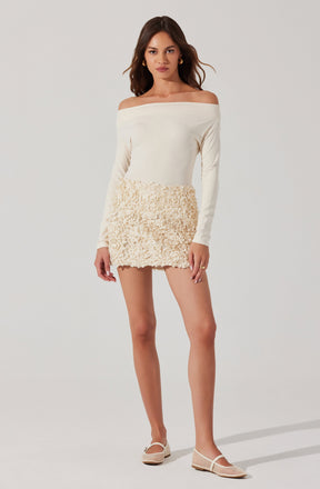 A woman is wearing an off-the-shoulder, long-sleeve white top paired with a cream-colored textured mini skirt. She has on white woven flats and gold hoop earrings.