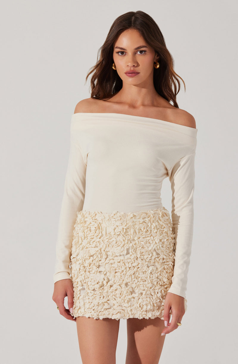 A woman is wearing an off-the-shoulder, long-sleeved, cream-colored top paired with a short, textured cream-colored skirt that has a ruffled, floral-like pattern throughout.
