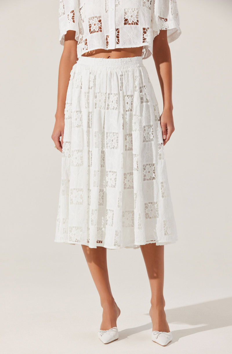 The image shows a woman wearing a matching white outfit made of lace fabric with a geometric square pattern. The top is a cropped, loose-fitting blouse with short sleeves, featuring the same lace pattern. The skirt is a high-waisted, midi-length A-line with an elastic waistband, made from the same lace material, creating a coordinated look. She is also wearing white pointed-toe high heels with decorative bows on the front.