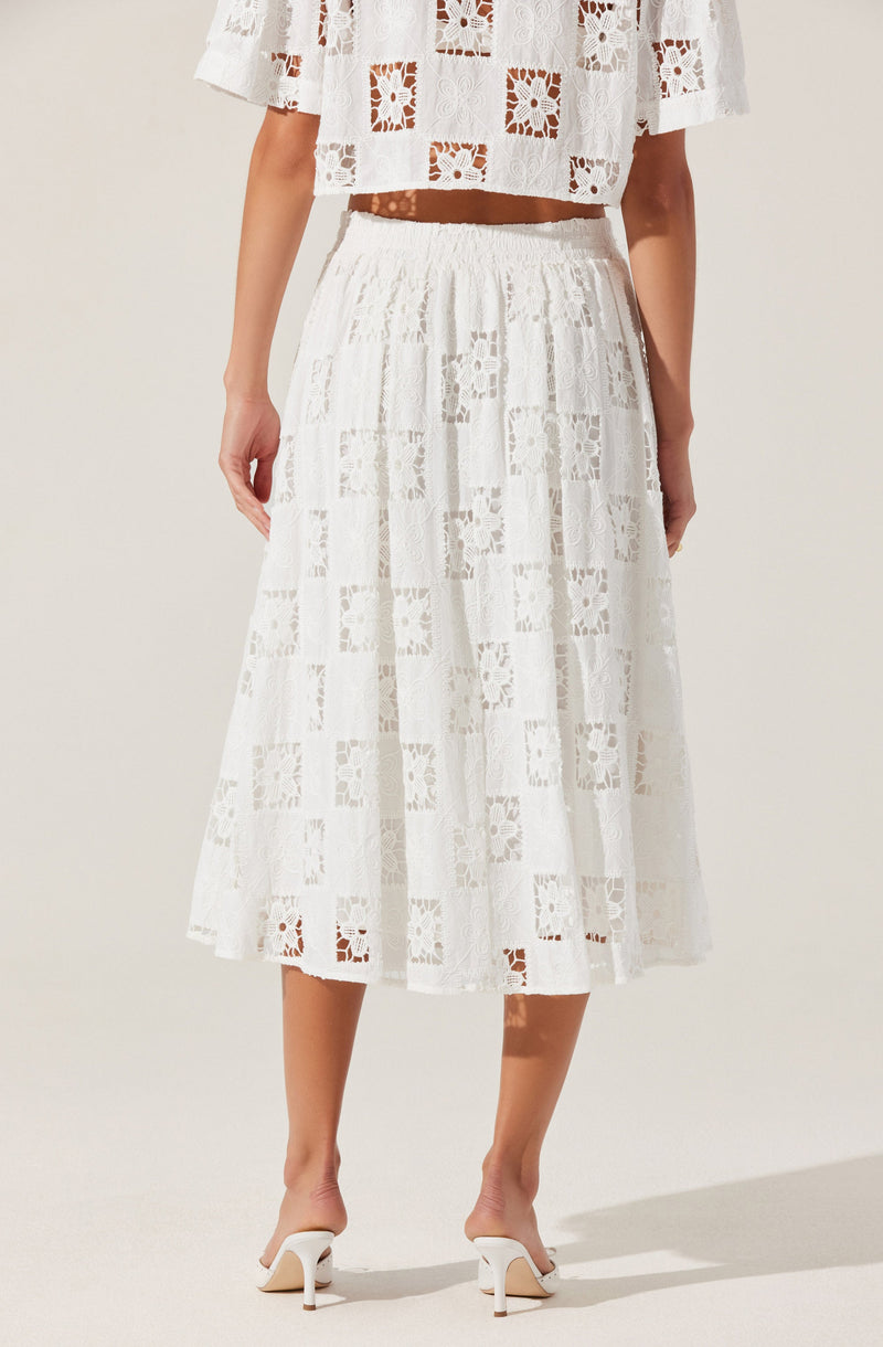 A woman is wearing a white, semi-sheer lace skirt with a patchwork pattern of squares and floral designs. The skirt has a high waist with an elastic band and falls to mid-calf length. She is also wearing white high-heeled shoes with ankle straps. The top she is wearing is a matching white lace off-the-shoulder crop top with a similar patchwork pattern.