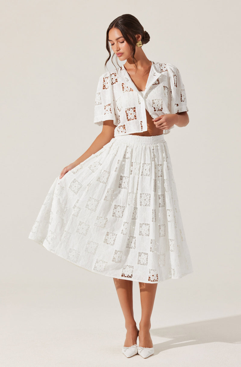 A woman is wearing a white two-piece outfit made of fabric with a lace or eyelet pattern. The top is a short-sleeved, button-up shirt with a collar, featuring square and floral cutouts. The skirt is a high-waisted, full-length A-line with a similar pattern of cutouts, giving it a textured, intricate appearance. She is also wearing white shoes with a pointed toe and a decorative bow.