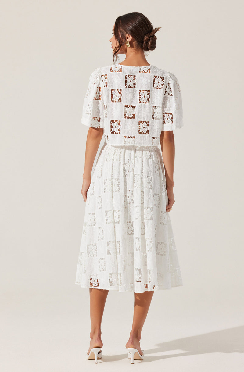 A woman is wearing a white, lace dress with a pattern of square and floral cutouts. The dress has a loose-fitting top with short sleeves and a high neckline, and a gathered, mid-length skirt that falls below the knees. She is also wearing white high-heeled shoes.