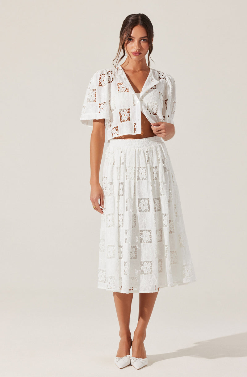 A woman is wearing a white, two-piece outfit made of lace with a pattern of squares and floral motifs. The top is a short-sleeved, button-up shirt with a collar, and the bottom is a high-waisted, midi-length skirt with an elastic waistband. She is also wearing white pointed-toe shoes with bows on the front.