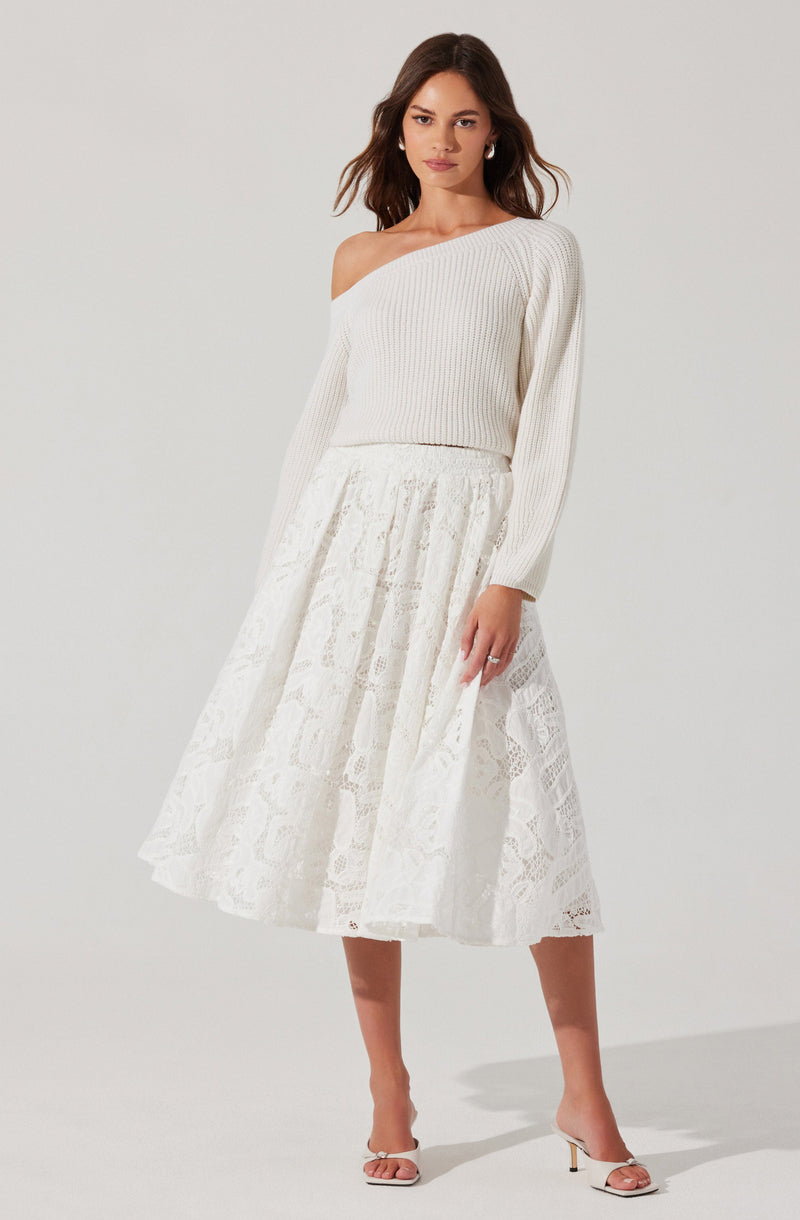 A woman is wearing an all-white outfit consisting of a loose, off-the-shoulder knit sweater with long sleeves, paired with a high-waisted, midi-length lace skirt featuring intricate patterns. She is also wearing white open-toe heels with ankle straps.