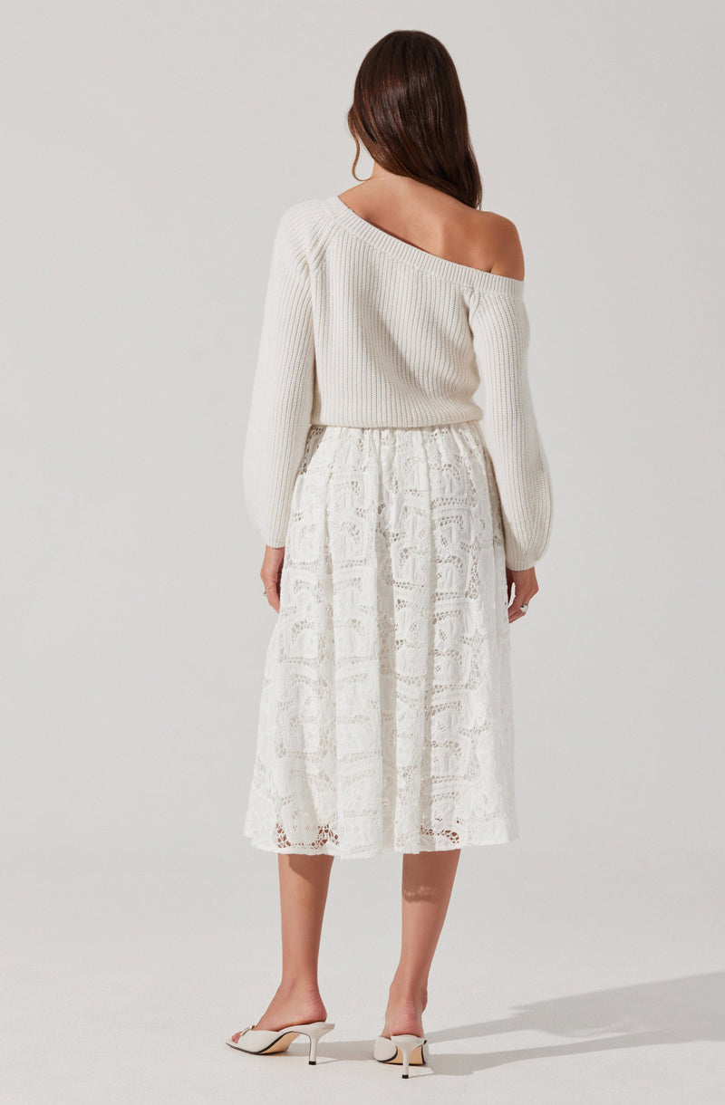 A woman is wearing an off-the-shoulder white knit sweater with long sleeves and a loose fit. She pairs it with a white, mid-length lace skirt featuring intricate patterns and a slightly gathered waist. She completes the outfit with white high-heeled shoes.