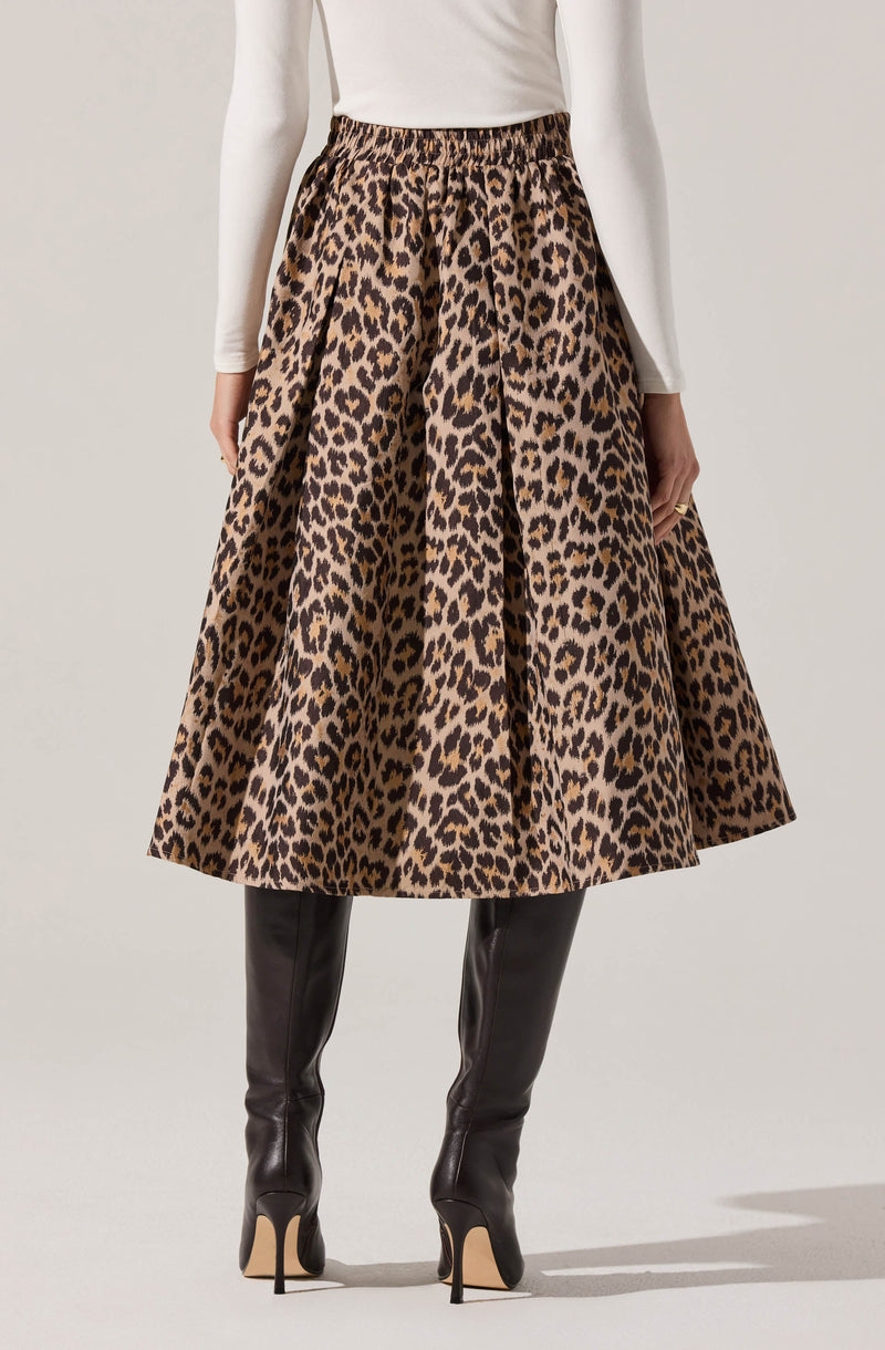 A woman is wearing a high-waisted, full-length skirt with a leopard print pattern in shades of brown and black. The skirt has a slight sheen and a voluminous, A-line silhouette. She pairs it with a long-sleeved, fitted white top and black knee-high boots with high stiletto heels.
