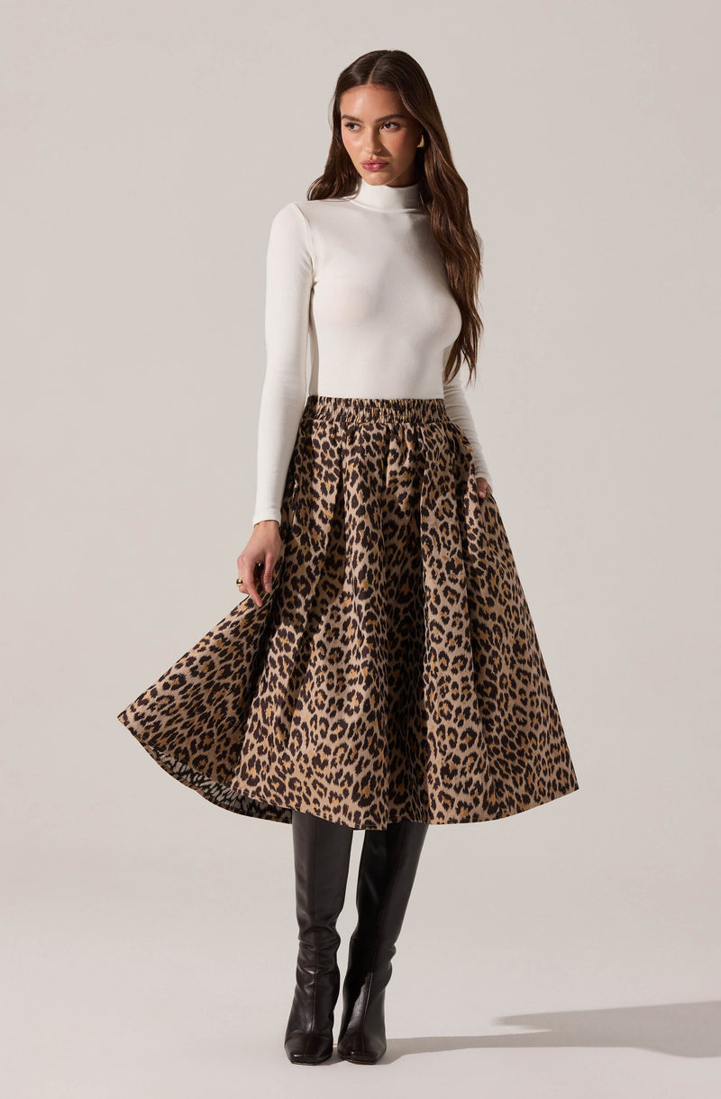A woman is wearing a fitted white long-sleeve turtleneck top paired with a high-waisted, ankle-length leopard print skirt that has a voluminous, flowing silhouette. She completes the outfit with black knee-high boots.