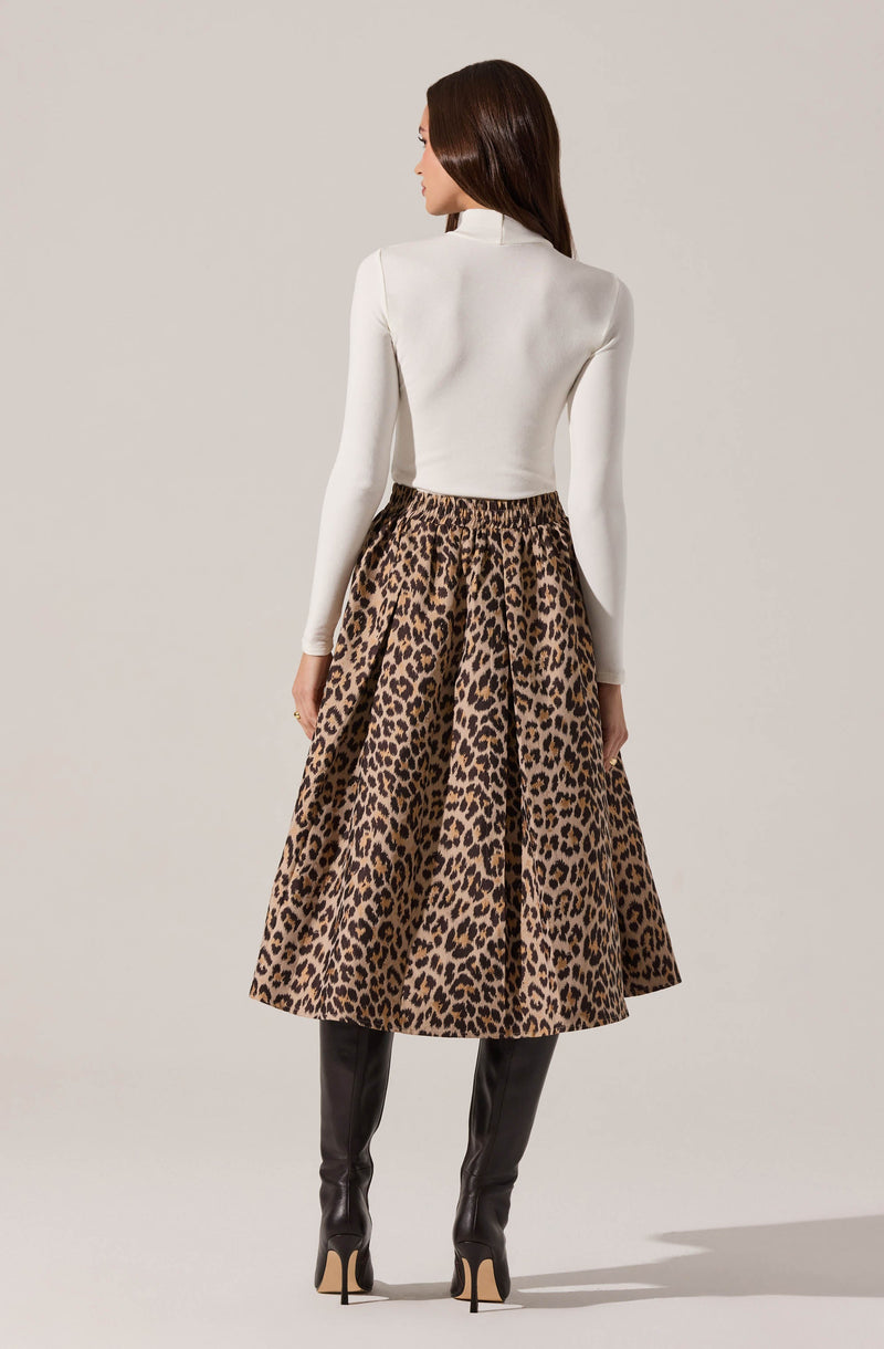 A woman is wearing a fitted white long-sleeve top and a high-waisted, full-length leopard print skirt with a voluminous silhouette. She also has on tall, black leather knee-high boots with high heels.