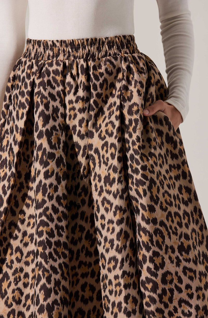 A person is wearing a high-waisted, loose-fitting skirt with a leopard print pattern in shades of brown, black, and beige. The skirt has an elastic waistband and is made of a flowing fabric. The person is also wearing a long-sleeved, light-colored top tucked into the skirt, with their hand resting in the side pocket.