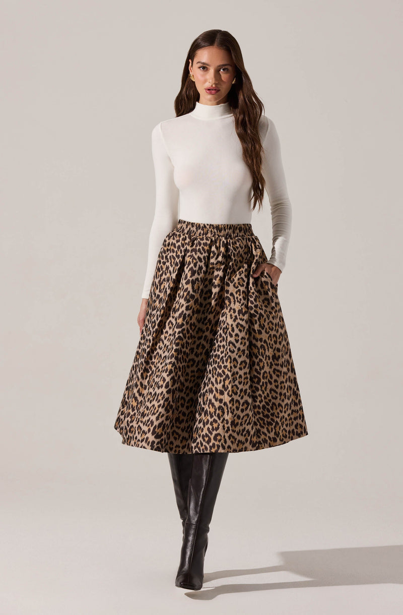 A woman is wearing a fitted, long-sleeved, white turtleneck top paired with a high-waisted, A-line skirt featuring a bold leopard print pattern in shades of brown and black. She completes her outfit with knee-high black leather boots.