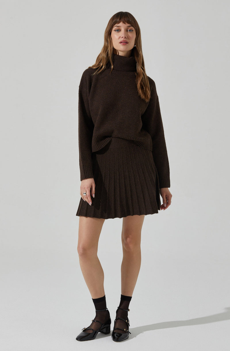 A woman is wearing a dark brown, oversized turtleneck sweater paired with a matching ribbed pleated skirt. She has on sheer black ankle socks and black pointed-toe shoes with a strap. The outfit appears cozy and stylish with a monochromatic brown color scheme.