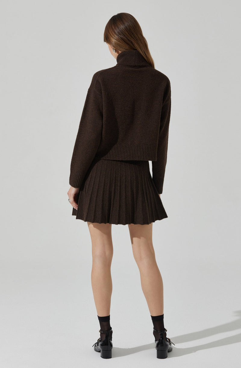 A woman is wearing a dark brown, oversized turtleneck sweater with long sleeves that extend past her wrists. She pairs it with a matching dark brown, high-waisted, pleated mini skirt that reaches mid-thigh. She completes the outfit with black ankle socks and black lace-up heels with block heels.