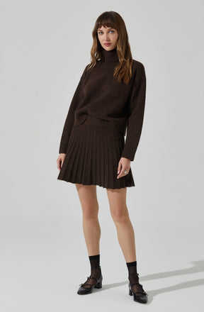 A woman is wearing a dark brown, long-sleeved turtleneck sweater paired with a matching pleated skirt. She has on black ankle socks and black heeled shoes.