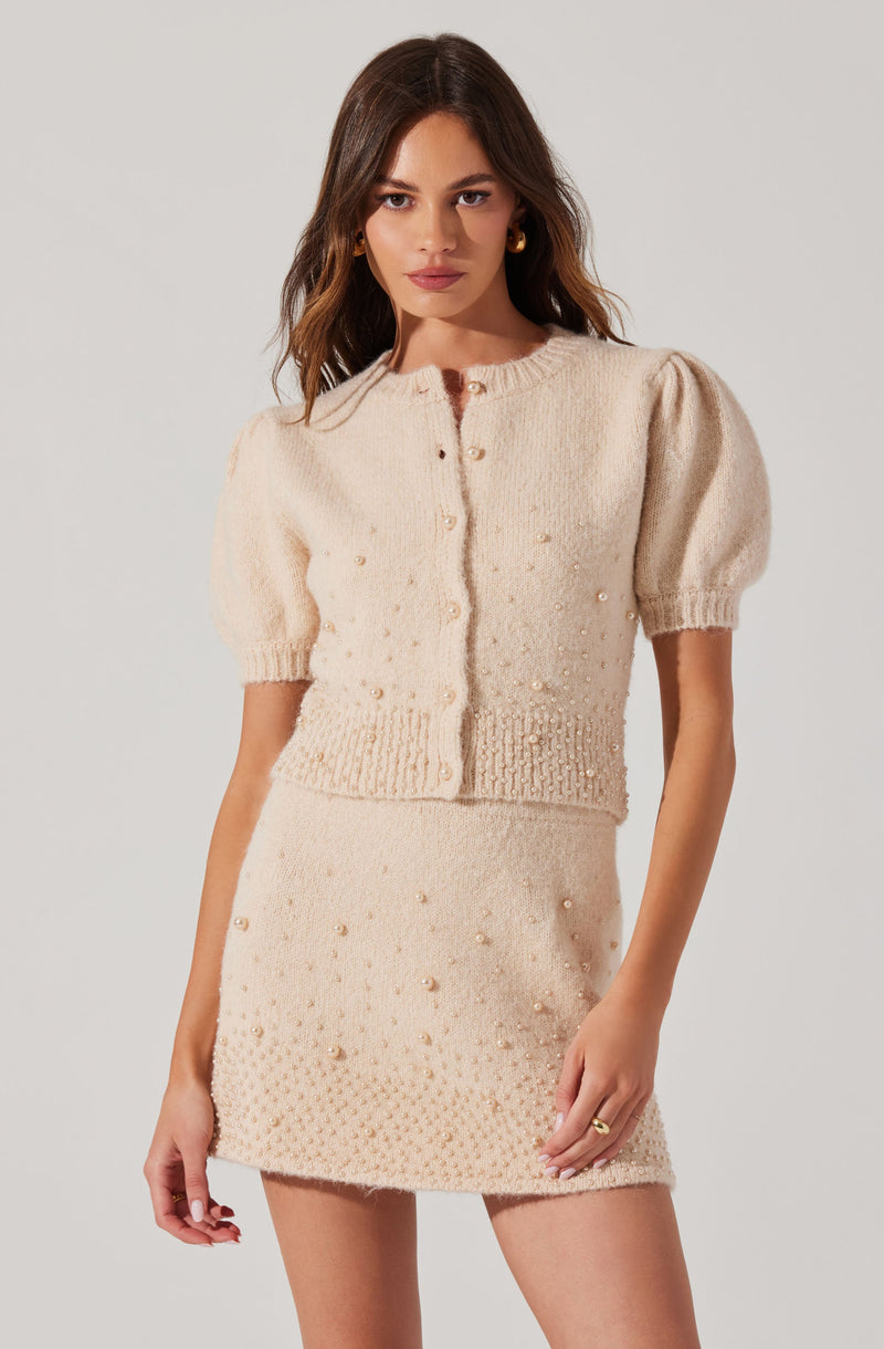 A woman is wearing a light cream-colored, knitted two-piece outfit. The top is a short-sleeved cardigan with a round neckline, buttoned down the front, featuring puffed sleeves and decorative pearl embellishments. The skirt is a matching mini skirt, also detailed with pearl embellishments, creating a coordinated, textured look.
