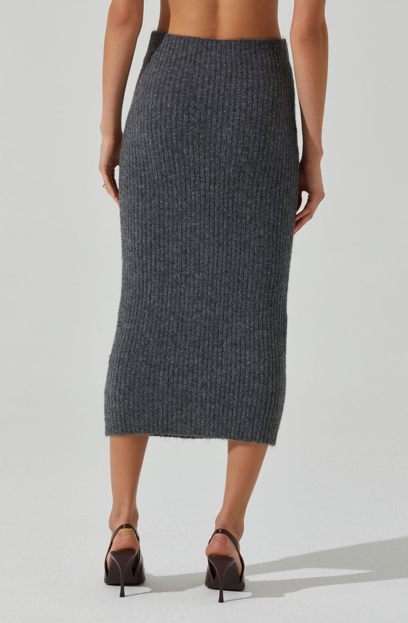 A woman is wearing a high-waisted, dark gray ribbed knit maxi skirt that extends down to her mid-calf. The skirt has a textured, vertical rib pattern and a fitted silhouette. She is also wearing black high-heeled shoes with ankle straps.