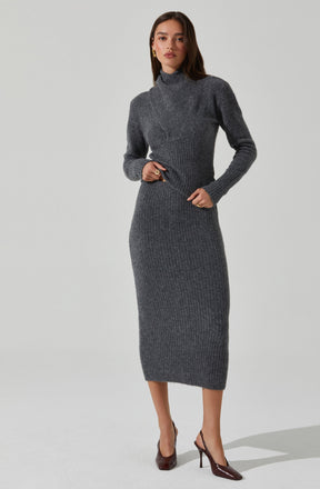 A woman is wearing a matching two-piece knit outfit consisting of a fitted, long-sleeved turtleneck top and a high-waisted, ankle-length pencil skirt. Both pieces are made of a dark gray, ribbed knitted fabric. She is also wearing pointed-toe high heels.