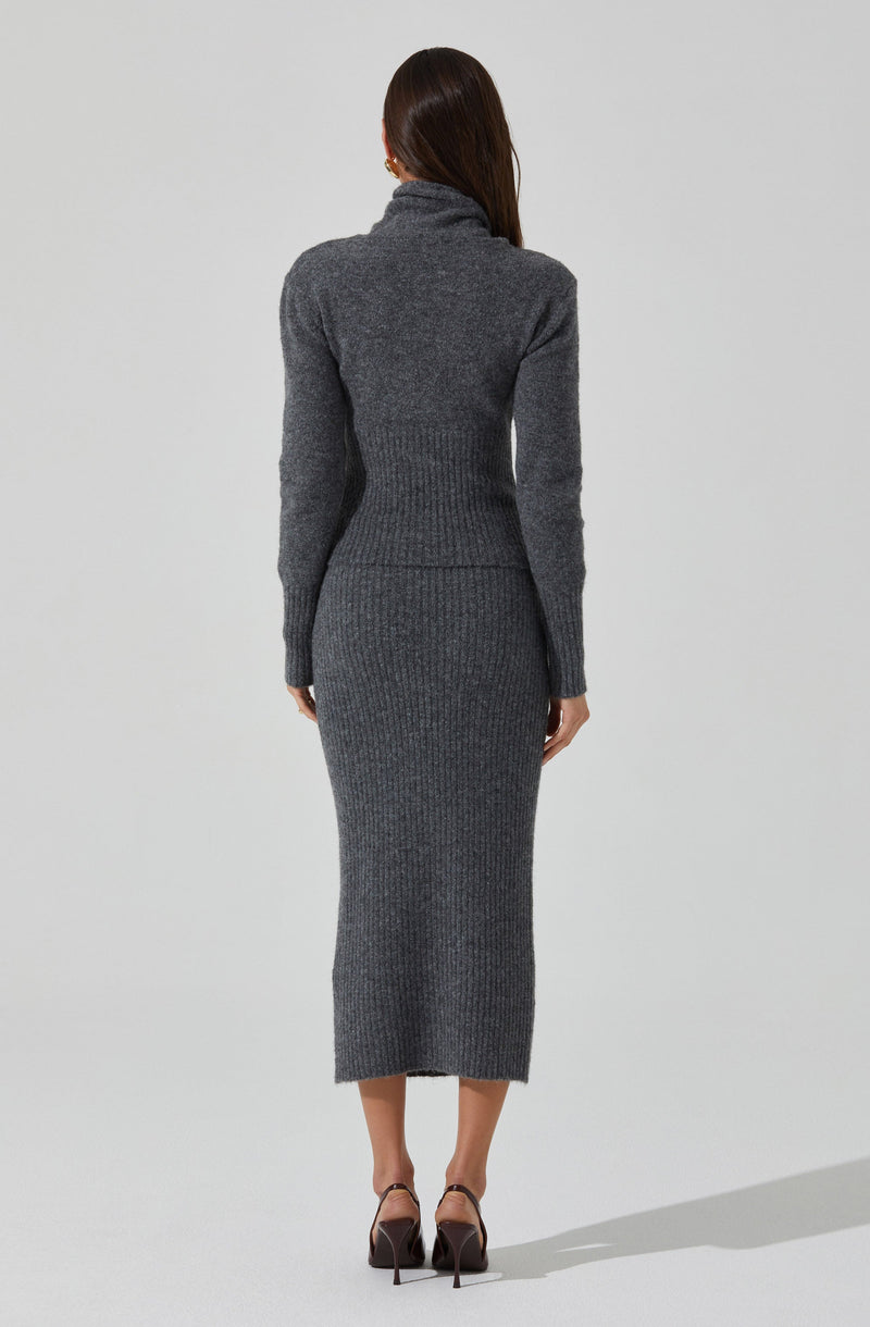 The image shows a woman wearing a matching dark gray cozy knit sweater and a long knitted skirt. The sweater has a high turtleneck collar and fitted long sleeves. The skirt is ribbed and extends down to her ankles. She is also wearing high-heeled shoes with ankle straps.