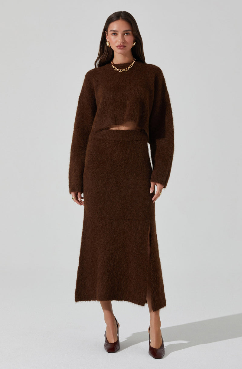 A woman is wearing a matching brown fuzzy knit set consisting of a long-sleeved cropped sweater and a high-waisted, ankle-length skirt with a side slit. She is accessorized with gold jewelry, including hoop earrings and a chunky chain necklace, and is wearing dark brown heels.