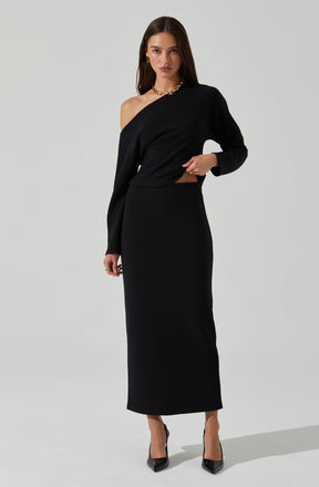 A woman is wearing a black, off-the-shoulder long-sleeve top paired with a high-waisted, full-length black skirt. She accessorizes with gold earrings, a gold necklace, and black high-heeled shoes.