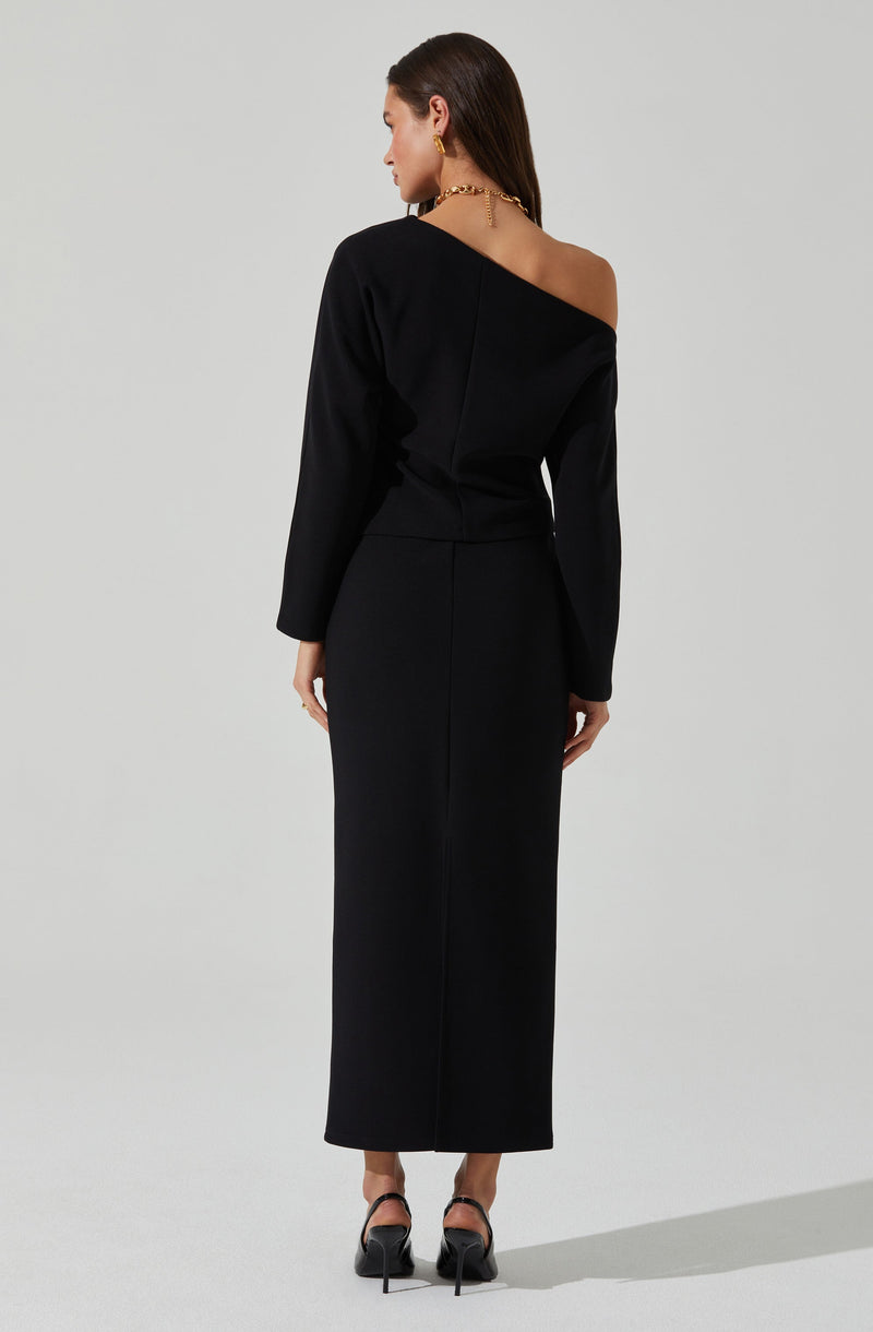 A woman is wearing a black, off-the-shoulder, long-sleeved top paired with a matching high-waisted, full-length skirt. She is also wearing black high heels with ankle straps. Her outfit is accessorized with gold jewelry, including earrings and a necklace.