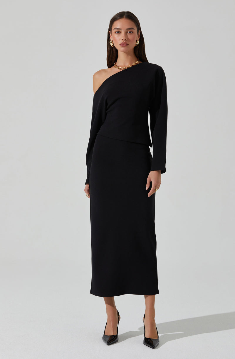 A woman is wearing a black, asymmetric, one-shoulder dress with long sleeves. She has accessorized with gold earrings, a gold necklace, and black pointed-toe heels.