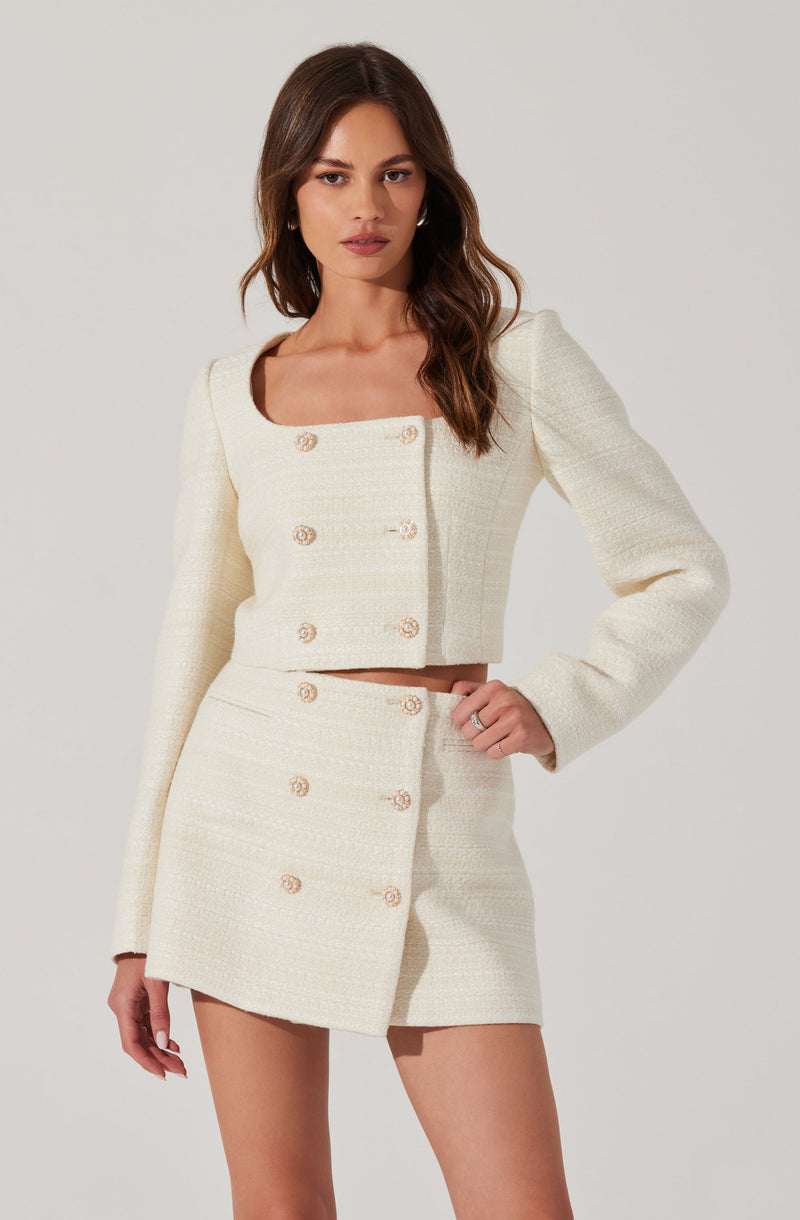 A woman is wearing a two-piece outfit made of cream-colored, textured fabric. The top is a cropped, long-sleeved jacket with a square neckline, featuring a double row of gold buttons down the front. The matching high-waisted skirt is short and has two rows of gold buttons running vertically along the front.