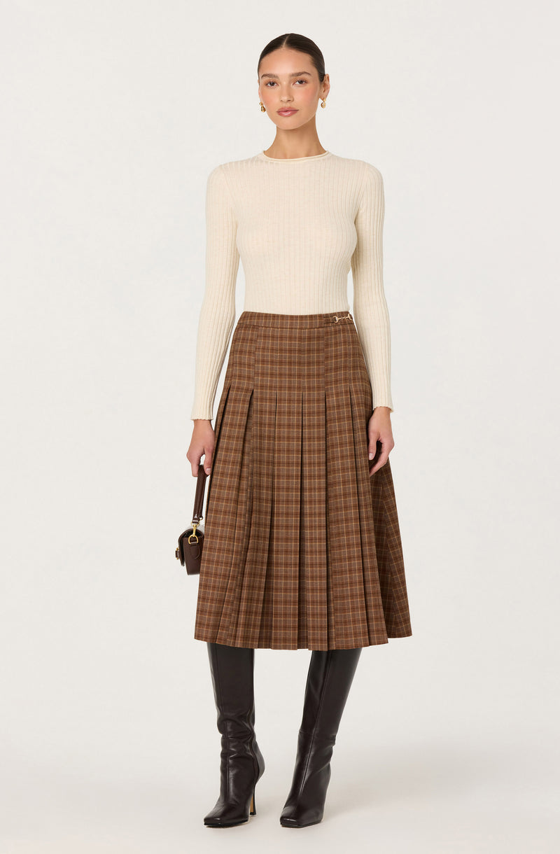 Woman wearing a cream ribbed long-sleeve top, a brown plaid pleated skirt, and black knee-high boots.