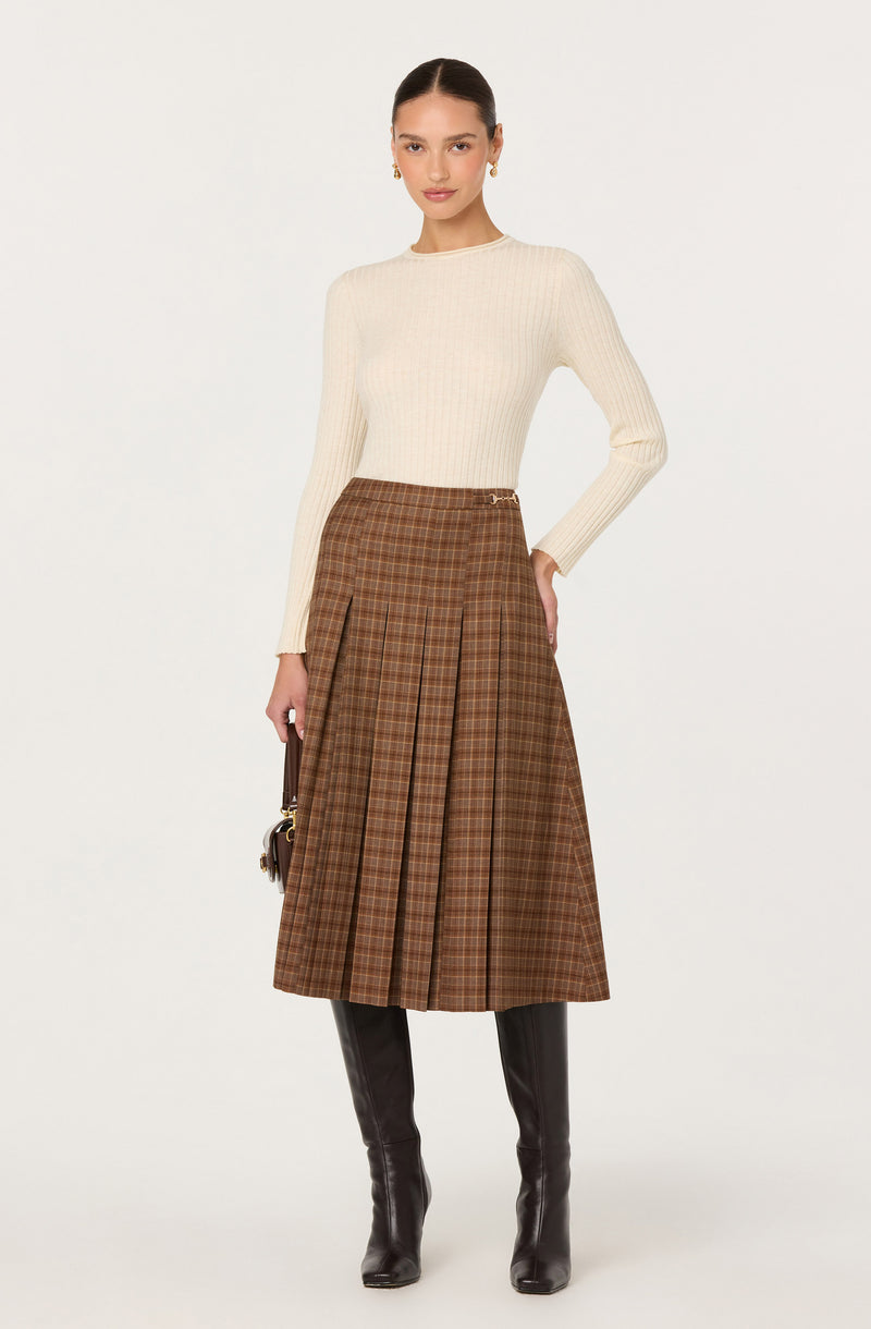 Woman wearing a cream ribbed sweater, brown plaid pleated skirt, and black knee-high boots.