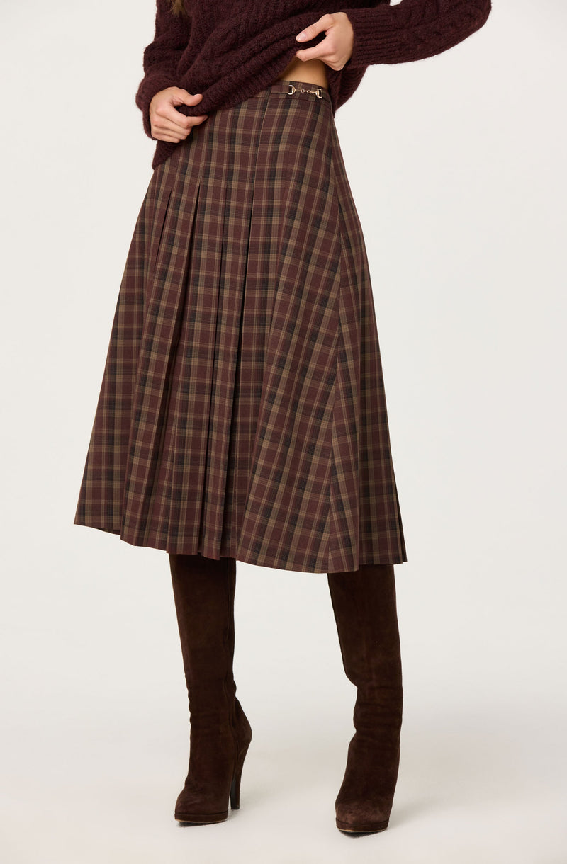 A woman is wearing a long, high-waisted, plaid skirt with pleats, featuring shades of brown, red, and green. She also has on a chunky maroon sweater and tall, dark brown suede boots.