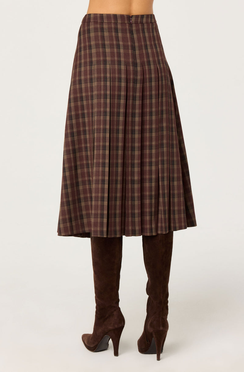 A woman is wearing a high-waisted, ankle-length skirt with a brown, red, and tan plaid pattern. The skirt features pleats at the hem and a zipper closure at the back. She is also wearing tall, dark brown suede high-heeled boots that extend above the knees.