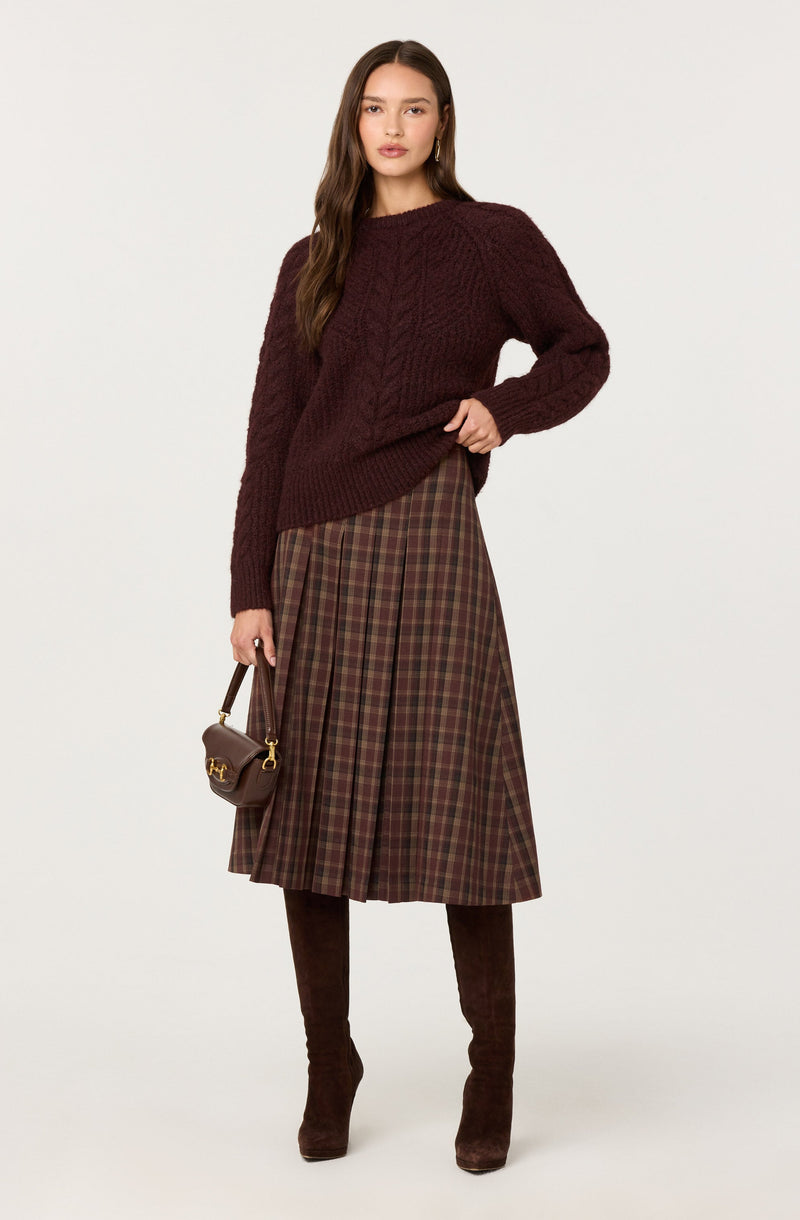 A woman is wearing a dark burgundy knit sweater with a cable pattern, paired with a high-waisted, pleated midi skirt featuring a tartan pattern in shades of brown and red. She is also wearing matching dark brown suede knee-high boots and carrying a small brown handbag with a gold clasp.