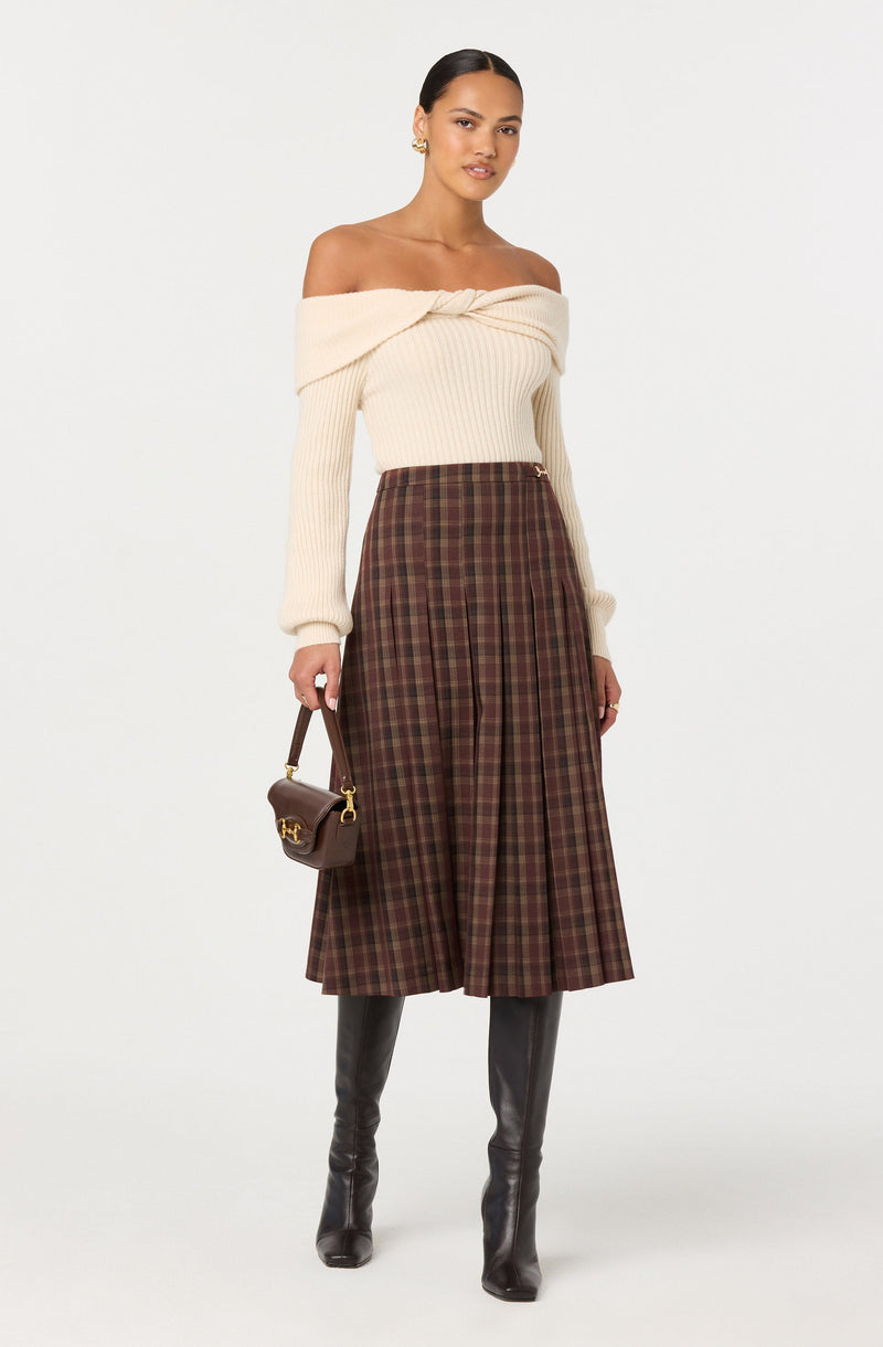 A woman is wearing an off-the-shoulder cream-colored knit sweater with a twisted detail at the neckline. She pairs it with a high-waisted, knee-length brown plaid skirt that has pleats. She is also wearing tall black leather boots and is holding a small brown handbag with gold hardware.