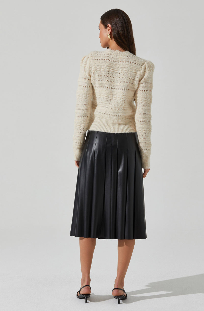 A woman is wearing a cream-colored, long-sleeved, knitted sweater with a textured pattern. The sweater has a fitted silhouette and slightly puffed shoulders. She pairs it with a black, high-waisted, pleated midi skirt made of a shiny material, and black open-toed heels with ankle straps.