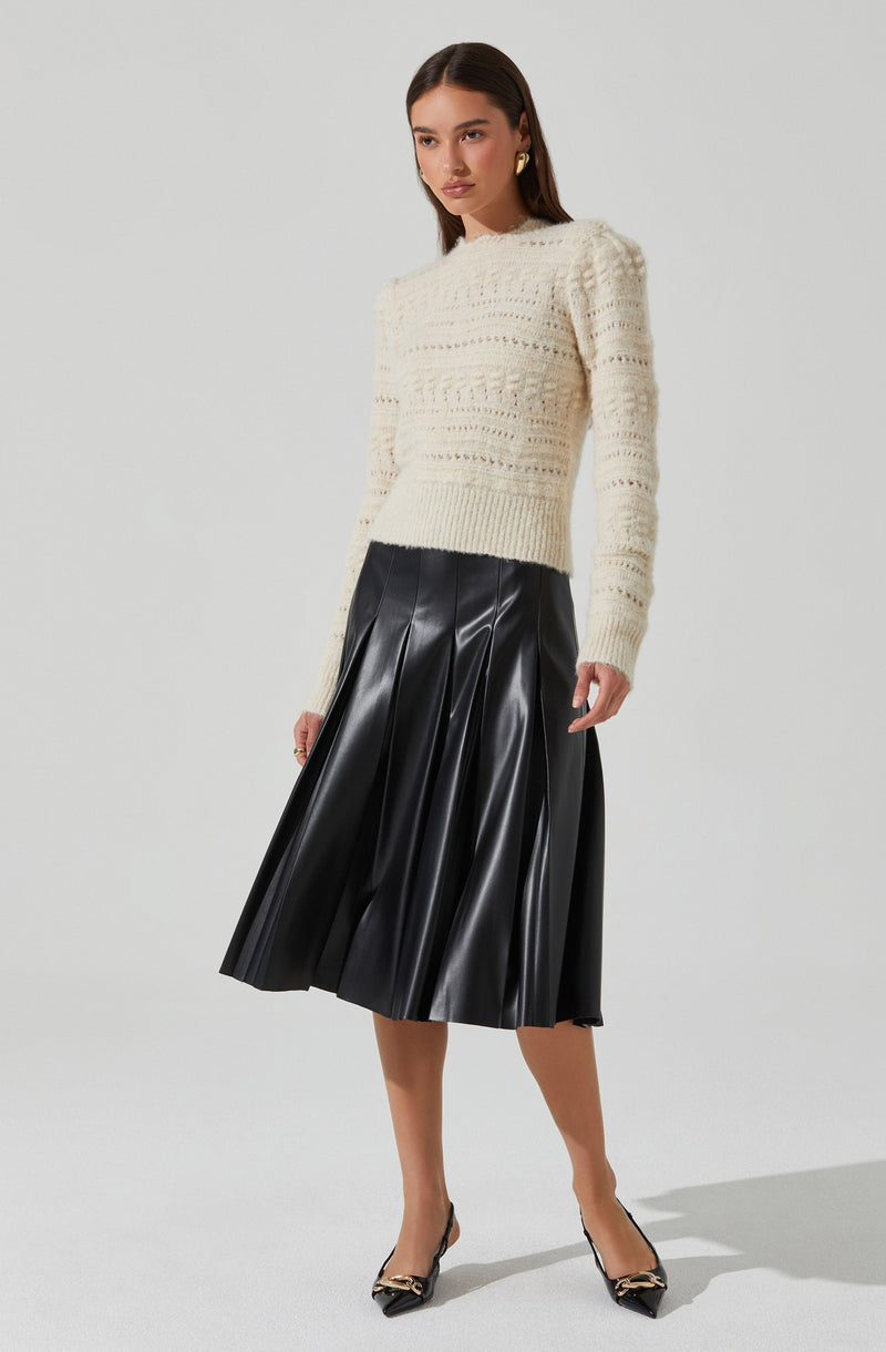 A woman is wearing a cream-colored, long-sleeved, knitted sweater with a textured, openwork pattern. She pairs it with a black, shiny, pleated midi skirt made of a leather-like material. She completes the outfit with black high-heeled shoes that feature a decorative gold element on the toe.